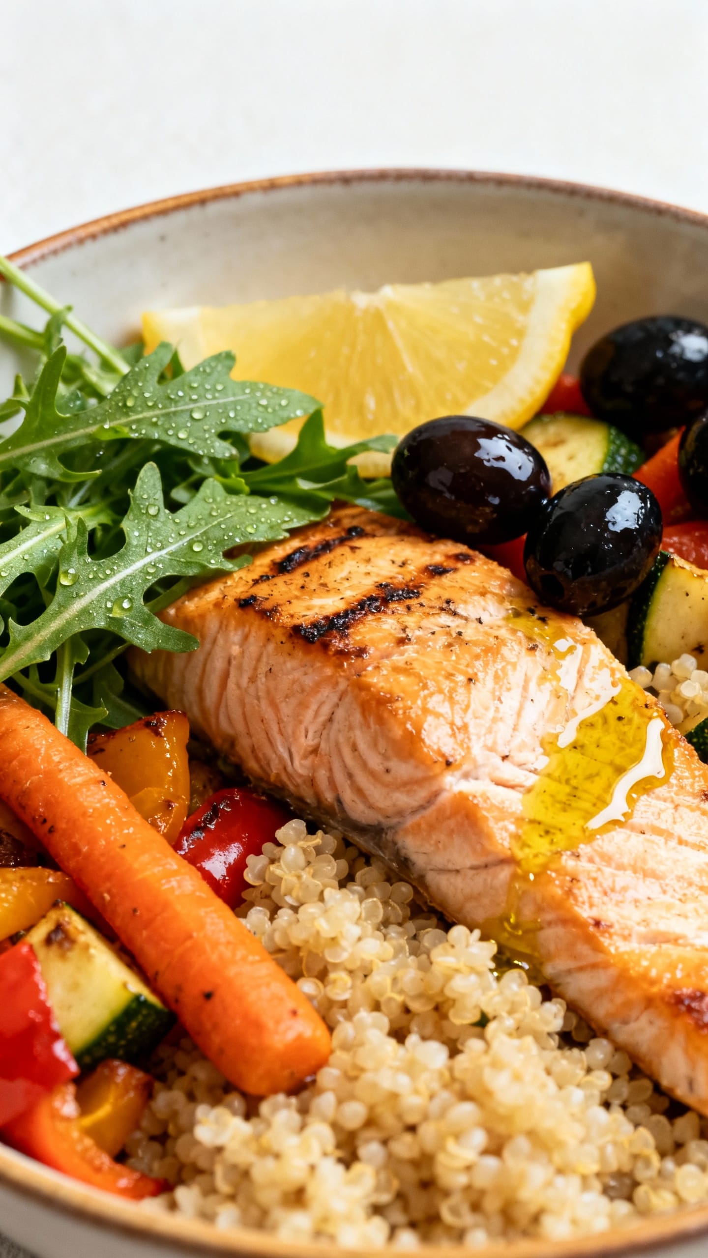 Closeup Mediterranean bowl: salmon, quinoa, roasted veggies, olives, arugula, lemon-olive oil drizzl