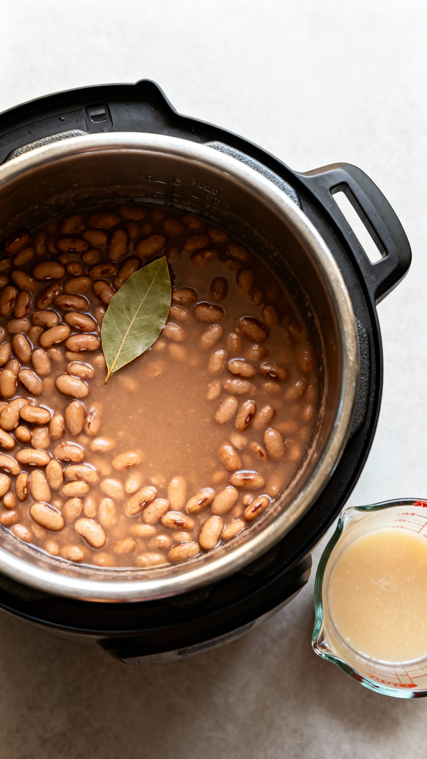 Overhead of Instant Pot with cooked pinto beans, bay leaf floating, reserved starchy cooking liquid